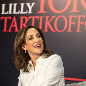 Close-up of Lilly Tartikoff at a book launch event. She wears a white blouse with bold red text on the backdrop behind her.