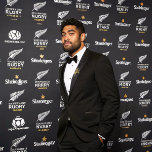Formal portrait of Akira Ioane in black tuxedo, standing at Steinlager Rugby Awards. Repeated logos and text on backdrop. Professional photo, focus on Ioane.