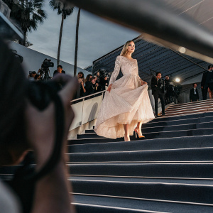Chiara Ferragni at Cannes Film Festival, wearing an elegant long-sleeved, off-white gown with lace detailing and a full skirt. She stands confidently on a bustling red carpet surrounded by flashing cameras and fans.