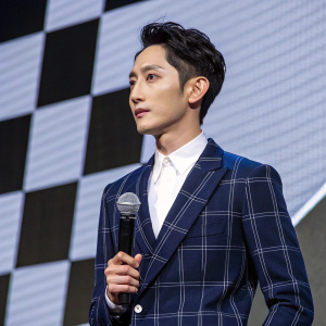 Lee Soo-hyuk, Korean actor, in a close-up shot. He is dressed in a navy blue plaid suit and crisp white shirt. Standing in front of a dynamic, checkered backdrop. Holding a microphone, he gives a determined look.