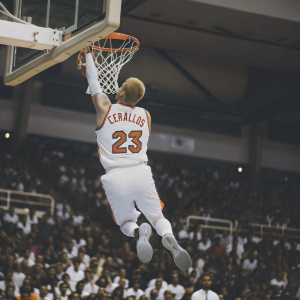 Chase Budinger mid-air during Slam Dunk Contest. Wearing white jersey with orange 'CERBALLOS' nameplate and number 23. Crowded arena with basketball hoop in background