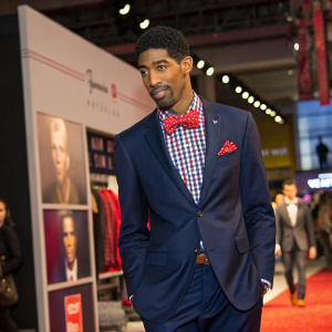 Fonzworth Bentley, African-American fashion icon, at a clothing launch event. Wearing a sharp navy suit with a checkered shirt and red bowtie. Poses confidently in front of an eye-catching billboard.