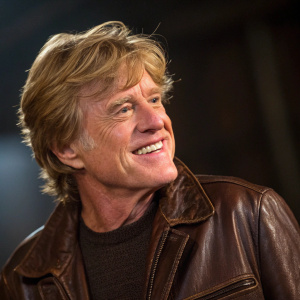 Close-up portrait of Robert Redford. Middle-aged, Caucasian man with long blonde hair. Wearing a brown leather jacket. Friendly smile. Dark, blurry background.