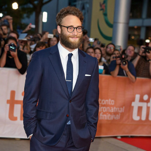 Seth Rogen at the Toronto International Film Festival. Bearded, wearing a tailored navy suit and tie. Surrounded by fans and flashing cameras
