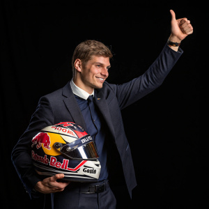 Max Verstappen, F1 driver, posing with a victorious thumbs-up. Dark suit and helmet, red and gold Red Bull logo. Stark black backdrop