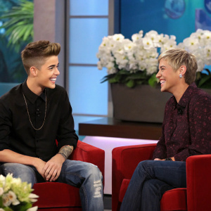 Justin Bieber and Ellen DeGeneres at a talk show. Justin in black shirt and blue jeans, Ellen in dark patterned shirt. Both smiling. Moderately lit indoor studio with colorful backdrop and white flowers.