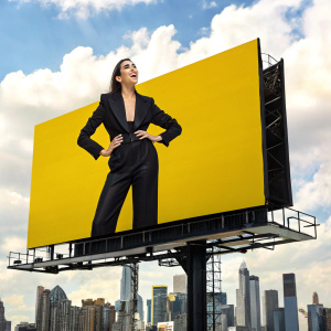Dua Lipa, in a sleek black outfit, stands confidently on a vibrant yellow background. She's posing for a billboard shot.