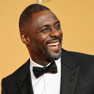Idris Elba, African actor, grinning. Wearing black suit, white shirt, black bow tie. Solid gold background at gala event.