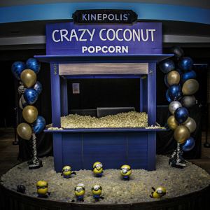 Promotional display for Kinepolis Crazy Coconut Popcorn at a movie theater. Minion-themed decorations featuring small figurines of minions in playful poses.