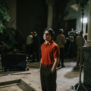Jake T. Austin, young actor, dressed in a red polo shirt. On set of a TV show, bustling crew in the background.
