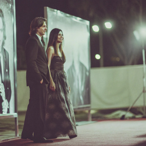 Dermot Mulroney and Tharita Cesaroni at a glamorous Hollywood premiere. Both dressed in elegant evening wear. Backlit by soft stage lights, with large movie poster backdrop