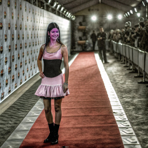 Charli D'Amelio, at the Nickelodeon Kids' Choice Awards 2023, stands confidently on a vibrant red carpet. She is dressed in a pink sleeveless dress with a black bodice and a ruffled skirt, against a backdrop filled with logos and graphics.