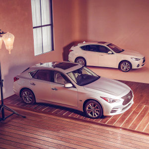 Digital illustration of Infiniti Q50 and Q30 car models. Q50 in front with sleek design, Q30 behind with compact design. Set against studio backdrop of warm golden hues.