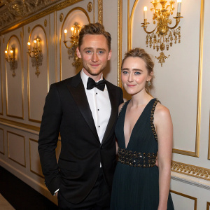 Tom Hiddleston and Saoirse Ronan, both in elegant attire, at a star-studded gala. Gilt-edged walls and chandeliers behind them.