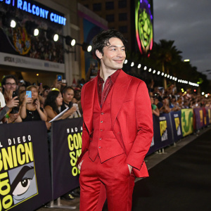 Ezra Miller at the San Diego Comic-Con, dressed in a vibrant red suit, standing in front of a crowd of fans. The background is filled with energetic cosplayers and convention banners.