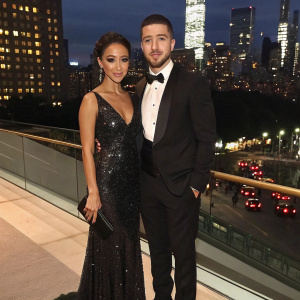 Melanie Iglesias and Vinny Guadagnino, standing together at the Met Gala. Melanie in a floor length sequined gown, Vinny in a tuxedo. Background is bustling New York City night skyline.