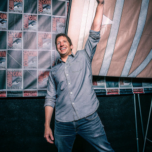 Andy Samberg at a Comic-Con event, dressed in a casual shirt and jeans. He's posing in front of a colorful, animated backdrop featuring Marvel and DC logos.