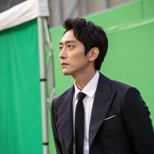 Lee Jun-Hyuk, actor, dressed in dark suit, white shirt, black tie. Standing in front of green screen, serious face