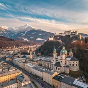 Aerial view of Salzburg, Austria, showcasing the picturesque city nestled among rolling hills. Snow-capped peaks, cobblestone streets, and iconic baroque buildings. The backdrop features a clear sky dotted with delicate clouds.