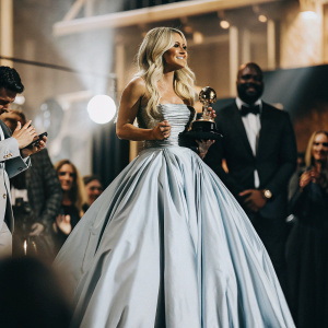 Carrie Underwood, blonde hair, in stunning silver gown. Accepting award at Grammys. Crowded room with flashing lights