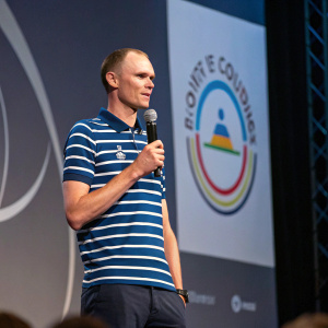 Chris Froome, in his 30s, with short hair, stands on a stage. Wearing a blue and white striped polo shirt. Addressing a crowd at a sports conference with a logo filled backdrop.