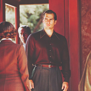 Henry Cavill, Caucasian male with intense gaze, in dark blue shirt and belt. Standing in front of blurred green background, surrounded by other individuals, natural lighting