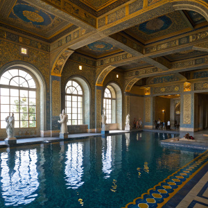 A majestic Roman-style pool at Hearst Castle in San Simeon, California. The water reflects ornate gold and blue patterns, arches, and large windows of the surrounding architecture. Statues of classical art dot the area. People admire the stunning details.