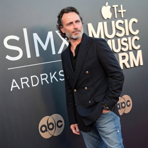 Andrew Lincoln, in a stylish black blazer and casual jeans, poses at the Apple Music Awards. SiriuxXM branded backdrop behind him
