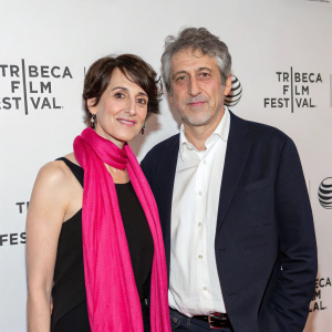 Dana Flor and Toby Oppenheimer attend the Tribeca Film Festival premiere. Dana, with short brunette hairstyle, dons a black sleeveless top paired with a vibrant pink scarf. Toby sports a neatly styled, graying haircut and wears a dark blazer over a white shirt. They stand against a sleek, minimalist backdrop.