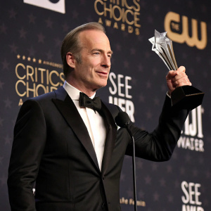 Bob Odenkirk accepting an award at the gala. Star-shaped trophy with shimmering details. Dark backdrop with event sponsors logos. Formal attire, black tuxedo with bow tie.