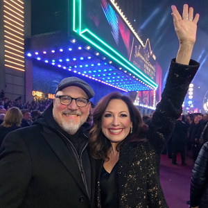 Frank Wildhorn and Linda Eder at a music award show, both smiling and waving to fans. Neon lights and crowded backdrop