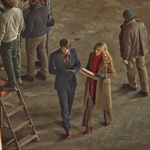 Chace Crawford and Blake Lively amidst bustling film set chaos. Crawford in a dark blue suit, focused on script. Lively in a brown coat and red scarf, displaying a look of concern. Other cast and crew visible in background. Candid photo capturing a moment of intense preparation.