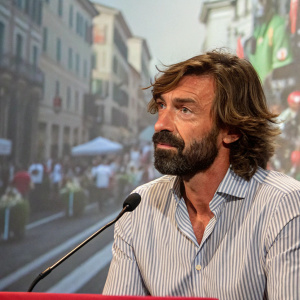 Former soccer star Andrea Pirlo, with a rugged beard and messy hair, wearing a casual striped shirt. At a press conference, behind him a bustling street scene.