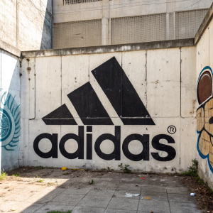 Graffiti art on concrete wall, featuring Adidas logo in bold, graphic style with strong contrast. Word 'adidas' written below in angular shapes.