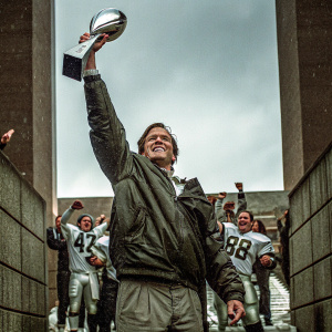 Tom Brady, in his New England Patriots gear, holding the Vince Lombardi Trophy high. Celebratory scene with team in background.