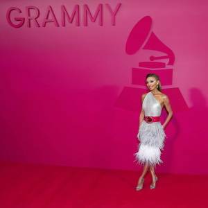 Giuliana Rancic on the Grammy red carpet. Wearing a sophisticated silver dress with a feathered skirt and red belt. Big Grammy logo backdrop