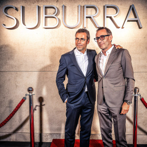 Luca Bianchini and Marco Bottinelli, both in suits, at the SUBURRA premiere. Luca is clean-shaven while Marco sports glasses and styled hair. The backdrop is a sleek, dark wall with the word SUBURRA in bold, elegant letters. Red carpet event.