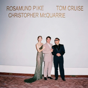 Rosamund Pike, Tom Cruise, and Christopher McQuarrie at a glamorous awards ceremony. Rosamund in a stunning emerald green gown, Tom in a sleek navy suit, and Christopher in a black tuxedo. The backdrop features the event logo.