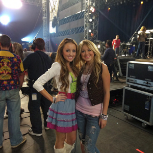 Miley Stewart and Lilly Truscott, from Hannah Montana, standing in a bustling backstage area at a concert venue.