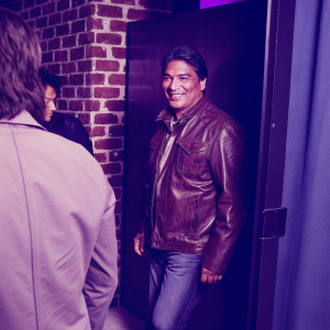 Gil Birmingham at the urban film festival. Brown leather jacket, casual attire. Warm smile, standing beside brick wall backdrop