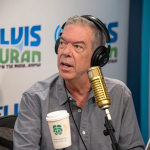 Elvis Duran, radio host, with intense look. Gray shirt, gold microphone. Starbucks coffee beside him. Branding backdrop in studio