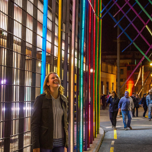 Jennifer Lawrence, smiling widely, wearing a casual outfit. A vibrant street scene behind her with people walking by.