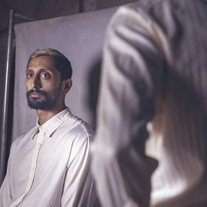 Riz Ahmed in a candid shot, sporting a beard and stylish haircut. Serious expression. Against a plain white backdrop.
