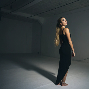 Kali Uchis, standing on a stark white background, wears a simple black dress. She poses with an air of mystery, her long hair cascading down her back. The setting is a quiet, dimly lit room.