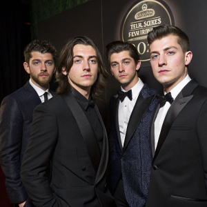 Arctic Monkeys band members Nick O'Malley, Jamie Cook, Alex Turner, and Matt Helders. Formal wear, posing stiffly. Dark background with intricate award show signage
