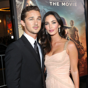 Shia LaBeouf and Megan Fox at the premiere party. Wearing a black suit and elegant gown respectively. Large movie poster backdrop featuring the film's title and artwork.
