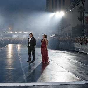 Rob Thomas and Marisol Maldonado, appearing at a Hollywood premiere. Rob in a black tuxedo, Marisol in a flowing red dress. Crowds and flashing cameras in the background.