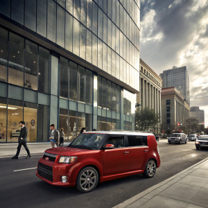 A vibrant red Scion xB parked on a bustling city street beside a sleek, modern office building.