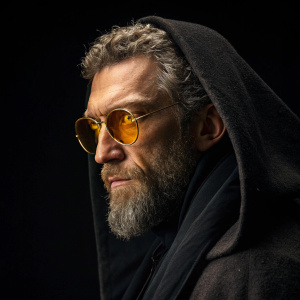 Vincent Cassel, with rugged beard and strong jawline, in dark cloak and glowing golden glasses. Dark and undefined background, making his features stand out sharply.