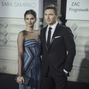 High-resolution close-up of Sara Sampaio and Zac Frognowski at a gala event. Sara in a stunning sequined gown with blue accents and a structured bodice, styled with contemporary hairstyle. Zac in a classic black tuxedo with a tie. White backdrop with event logos and sponsor names.
